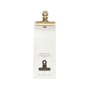 Guldklips fra Monograph med skinnende finish, hul til ophæng, fastgjort til emballage med størrelsesangivelse.