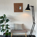 Rustik korktavle på væg, bænk med pude under, plante ved siden af, sort gulvlampe til højre.