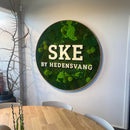 Rundt vægdekoration med teksten "SKE BY HEDENSVANG" på grøn baggrund, ved siden af et bord.