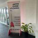 Stående displaybanner, rød/hvid design, tekst "AKTIV OVERVÅGNING", ikoner, kurvede standfødder, plante.