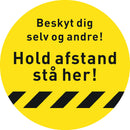Gul cirkulær gulvsticker med sort tekst: "Beskyt dig selv og andre! Hold afstand stå her!"