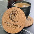 Runde korkbrikker med "COMPASSION HOUSE" logo, placeret på mørk overflade ved siden af kaffekop.