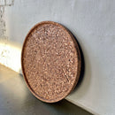 Rundt produkt med kant og tekstur, lænet mod hvid væg på hårdt underlag.