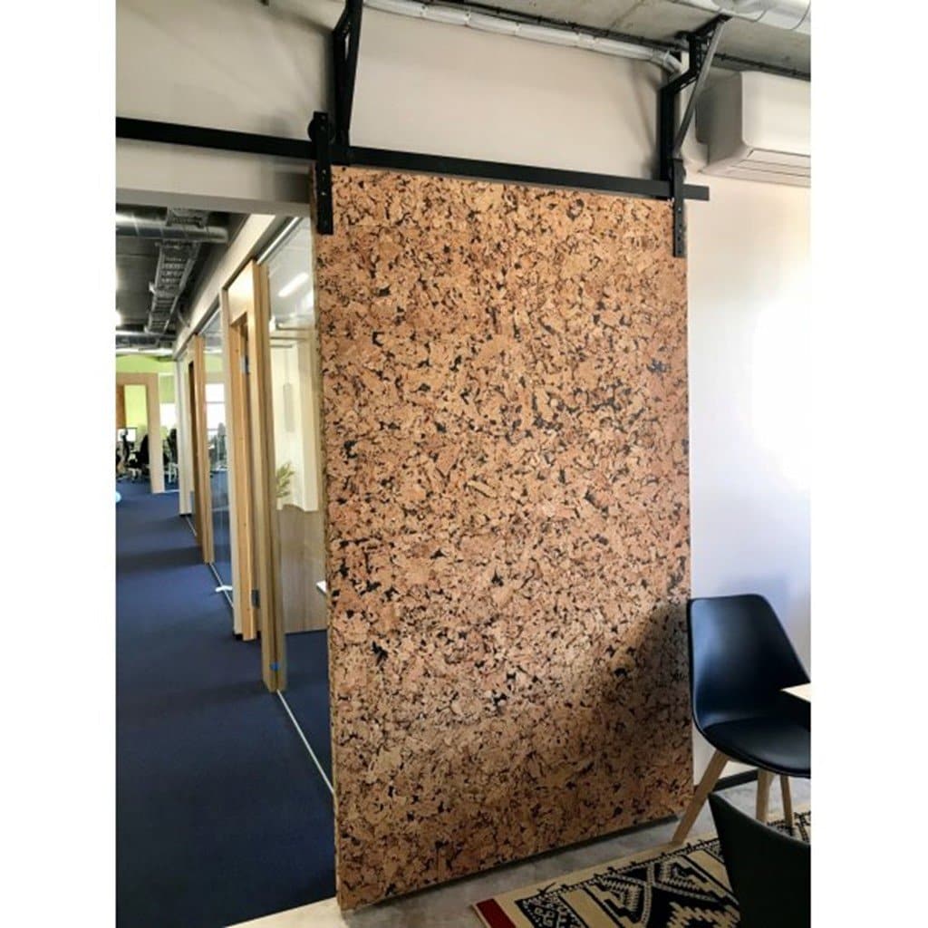 Stor, specklet korkpanel i døråbning, monteret på skinne. Stol og borddel synlige ved siden af.